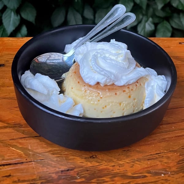 Bien Y Tu flan dessert with whipped cream and two spoons in a black bowl on a wooden table.