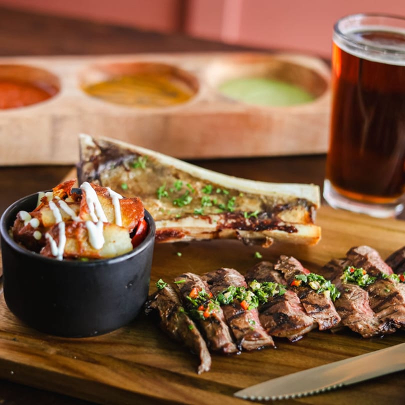 Mexican grilled steak, bone marrow, potatoes, and beer on a wooden board with sauces.