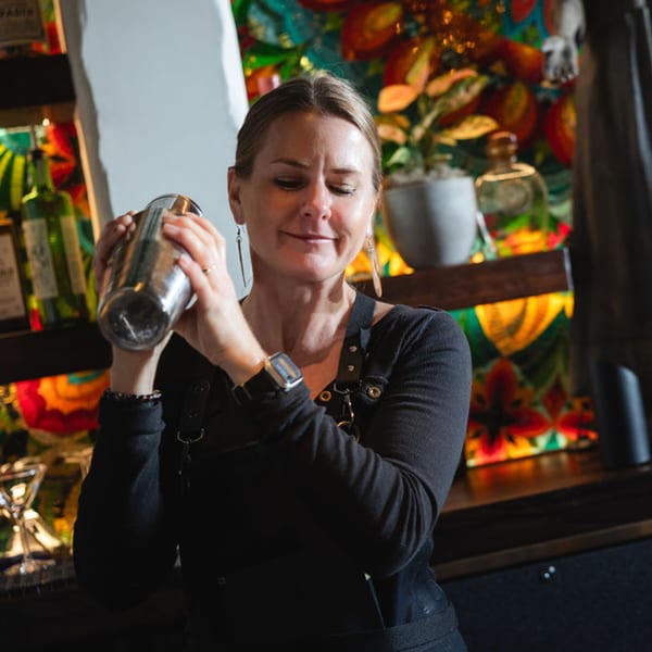 A female bartender at Bien Y Tu shakes a cocktail shaker with a colorful Mexican-themed background.