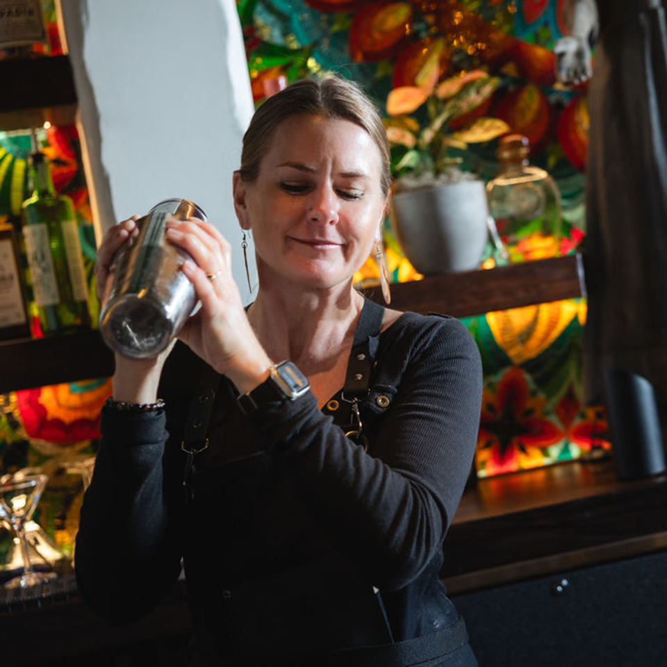 A female bartender at Bien Y Tu shakes a cocktail shaker with a colorful Mexican-themed background.
