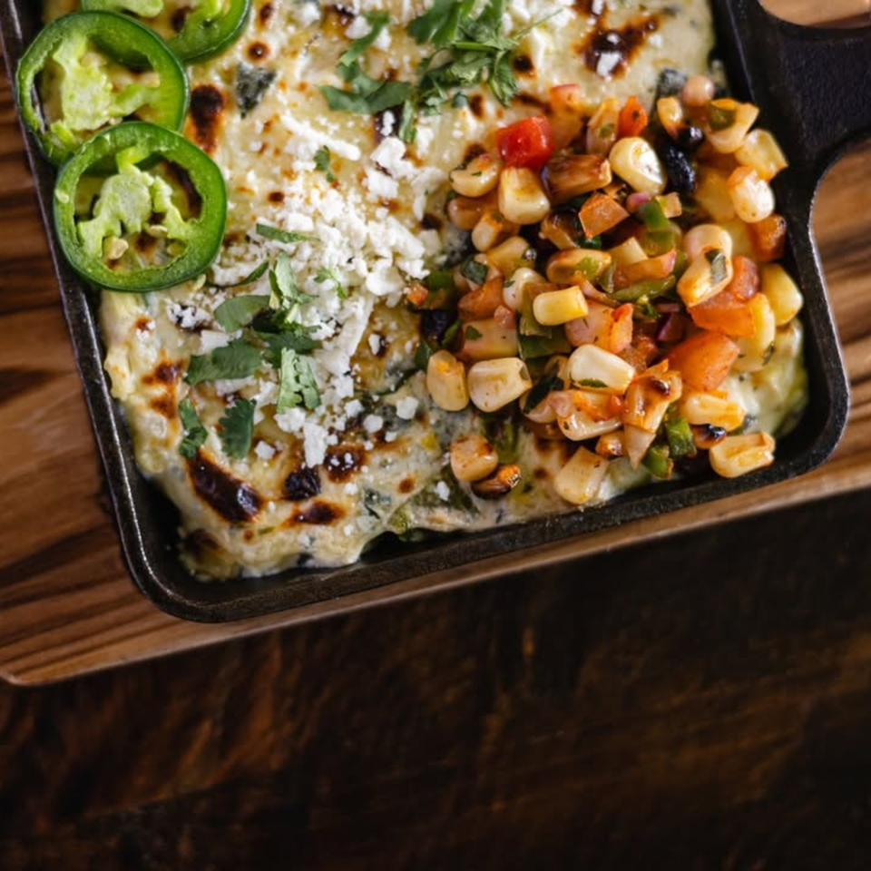 Bien Y Tu's Mexican esquites in a cast iron skillet, topped with cheese, cilantro, and jalapeños.