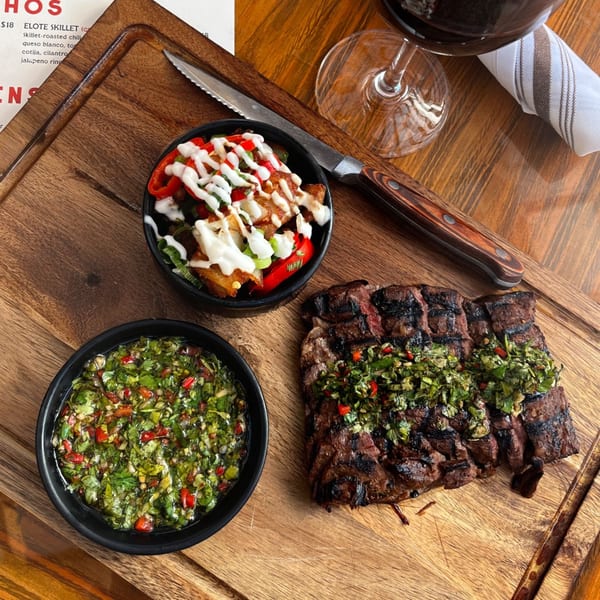 Grilled steak with chimichurri, a side of elote skillet, and a bowl of green salsa on a wooden board.