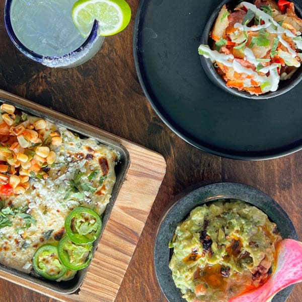 Overhead view of Mexican dishes: a margarita, cheesy corn dip, loaded nachos, and guacamole.
