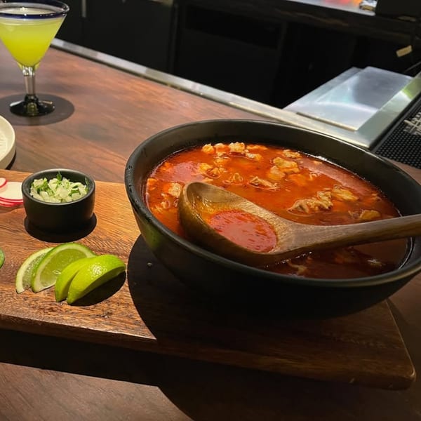 A bowl of red Mexican pozole soup with a wooden spoon, limes, and a green drink.